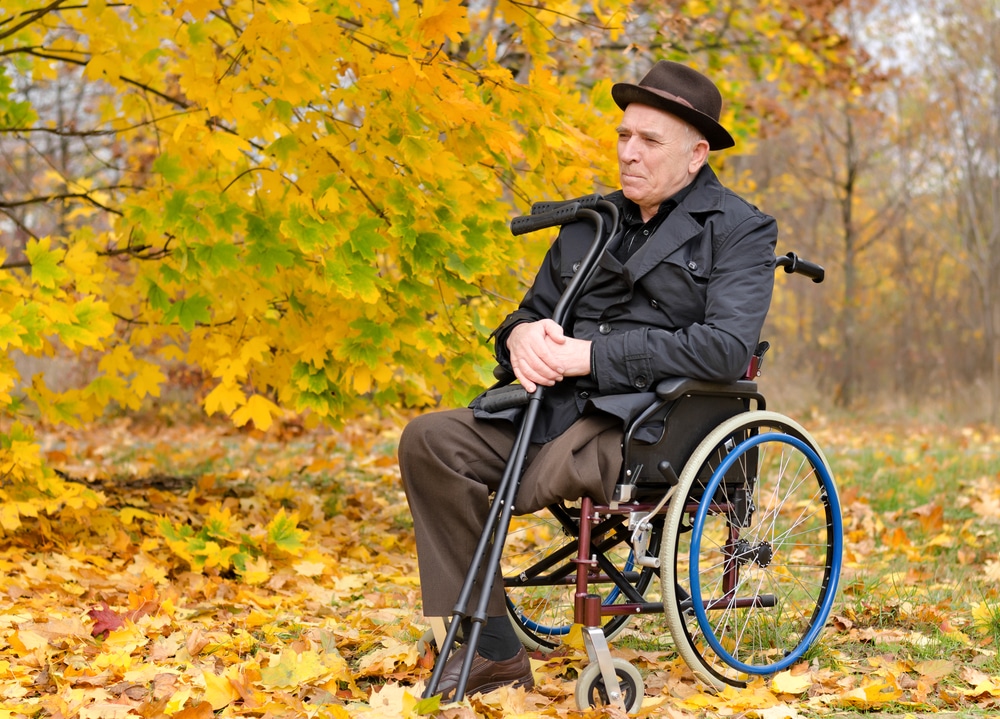 Thoughtful senior amputee sitting outdoors in his wheelchair with his crutches in his hand enjoying the last warmth of the autumn sun against a backdrop of colourful yellow leaves